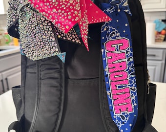 Peut inclure: Un sac à dos noir orné d'un grand nœud de cheerleading rose vif couvert de strass sur la bretelle gauche et d'une bande de tissu bleu à motif bandana sur la bretelle droite, portant le nom CAROLINE en lettres roses scintillantes.