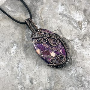 May include: A handcrafted pendant featuring a purple and white stone, intricately wrapped in dark wire. The oval-shaped stone has a marbled appearance. The pendant hangs from a black cord, showcasing a unique, artistic design.