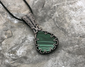 May include: A handcrafted pendant featuring a teardrop-shaped malachite stone, encased in a wire-wrapped setting. The pendant hangs from a black cord. The malachite displays a rich green color with distinctive banding.