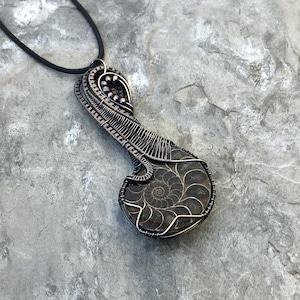 May include: A handcrafted pendant featuring a fossilized ammonite encased in intricate wire wrapping. The pendant showcases a dark, spiral-patterned fossil, suspended from a black cord. The wirework is a dark, metallic color, adding a unique, artistic touch.