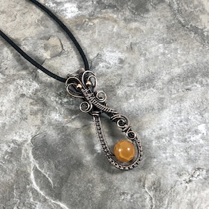 May include: A pendant necklace featuring a round, amber-colored stone encased in a dark, oxidized copper wire-wrapped frame. The wire design includes intricate swirls and loops at the top, suspended from a simple black cord necklace.
