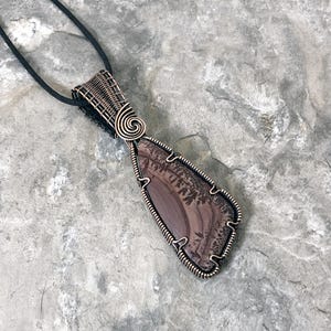 May include: A copper wire wrapped pendant featuring a brown and white agate stone with a dendritic pattern. The pendant is attached to a black cord.