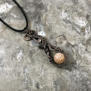 May include: A handcrafted copper wire pendant necklace with a light brown, textured stone bead. The pendant has intricate wire wrapping and a small, clear crystal accent. The necklace hangs on a black cord.