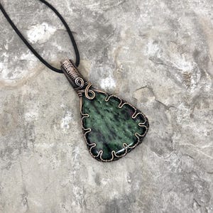 May include: A green and black gemstone pendant with copper wire wrapping. The pendant is hanging from a black cord.