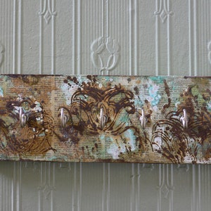 Könnte beinhalten: Ein rechteckiger Schlüsselhalter zur Wandmontage mit vier silbernen Haken. Die Holzplatte hat ein Used-Design mit braunen, türkisfarbenen und weißen Farbspritzern über einem Vintage-Zeitungsdruck. Dekorativer, rustikaler Stil.