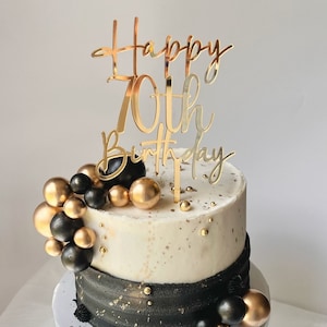 May include: A white and black cake with gold sprinkles and a gold 'Happy 70th Birthday' topper. The cake is decorated with black and gold round ornaments.