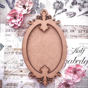 May include: A decorative wooden oval frame with ornate detailing, set against a backdrop of vintage paper with floral and script elements. Pink paper flowers are arranged around the frame, creating a romantic, vintage aesthetic.