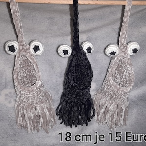 Könnte beinhalten: Drei gehäkelte Hängeschmuckstücke, zwei in Beige und eines in Schwarz, jedes mit einem Paar schwarzer Augen und einer Fransen an der Unterseite. Der Text "18 cm je 15 Euro" ist am unteren Rand des Bildes sichtbar.