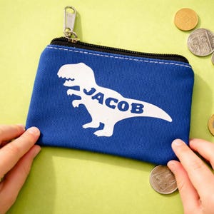 Op de afbeelding: Een rechthoekige blauwe canvas portemonnee met een witte silhouet van een Tyrannosaurus Rex, waarbij de naam JACOB in blauwe letters in het midden van de dinosaurus is gedrukt. De portemonnee heeft een zwarte ritssluiting met een zilverkleurige metalen trekker.