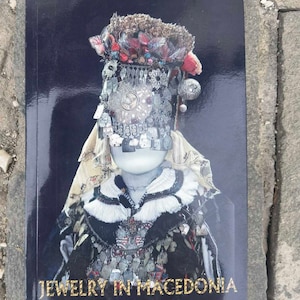 May include: A book cover featuring a photograph of a person adorned in traditional Macedonian jewellery. The jewellery is silver and boasts intricate designs. The book title is "Jewelry in Macedonia".