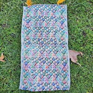 May include: A white fabric cushion with a colorful geometric pattern and yellow tassels on each end. The cushion is lying on a bed of green grass.