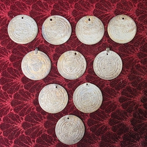 Puede incluir: Una colección de diez monedas antiguas de plata con intrincados diseños en relieve. Cada moneda redonda tiene un pequeño agujero en la parte superior, posiblemente para usar como colgante. Las monedas están dispuestas sobre una tela roja oscura texturizada.