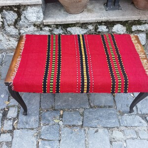 Könnte beinhalten: Ein roter gewebter Teppich mit schwarzen, grünen und gelben Streifen liegt auf einer Holzbank mit schwarzen Beinen. Die Bank steht auf einem Kopfsteinpflaster.