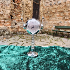 Puede incluir: Un reloj plateado con una esfera redonda y un soporte de muelle. El reloj tiene dos antenas en la parte superior y está sentado sobre una superficie de terciopelo verde.