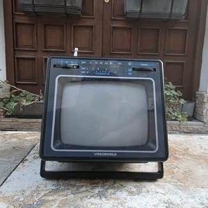 Könnte beinhalten: Ein alter, tragbarer Fernseher in Schwarz und Silber mit dem Wort "VIDEOMOBILE" auf der Vorderseite. Der Fernseher hat einen kleinen Bildschirm, einen Drehregler und einen eingebauten Ständer. Der Fernseher steht auf einer Betonoberfläche.