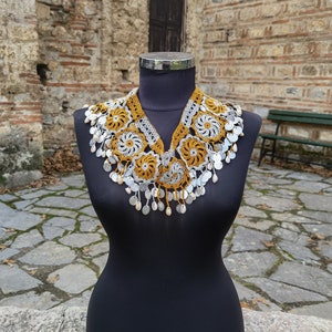 May include: A yellow and white crocheted collar with silver beads hanging from the bottom. The collar is displayed on a black mannequin against a stone wall background.