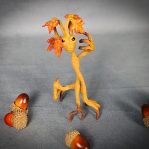 Peut inclure: Une petite figurine d'arbre humanoïde de couleur beige avec des feuilles d'érable orange et jaune sur la tête, debout sur de fines jambes en forme de racines. La figurine a des yeux sombres et est entourée de trois glands bruns.