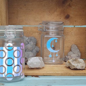 Peut inclure: Deux bocaux en verre transparent avec couvercles à charnière. L'un présente un motif de cercles irisés, l'autre un croissant de lune bleu et des étoiles. Les bocaux sont sur une étagère en bois avec des pierres décoratives.