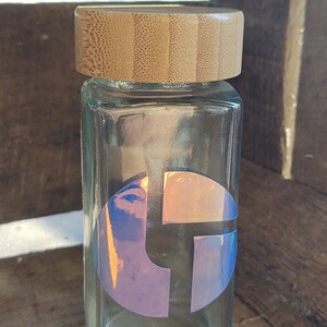 Op de afbeelding: Een glazen pot met een deksel van bamboe. De pot heeft een ronde sticker met een holografisch ontwerp van een gestileerde letter "L" in een cirkel.