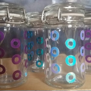 Peut inclure: Quatre bocaux en verre avec des couvercles en métal et un motif géométrique coloré de cercles violets et bleus. Les bocaux sont transparents et ont un aspect vintage.