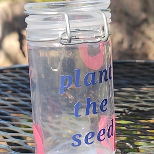 Peut inclure: Un pot en verre transparent avec un couvercle argenté et un motif rose et bleu. Le pot porte l'inscription "plant the seed".