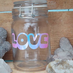 Peut inclure: Un bocal en verre transparent avec un couvercle à charnière et un fermoir argenté. Le mot "LOVE" est affiché sur le devant dans une police holographique multicolore. Le bocal est vide et repose sur un fond en bois.