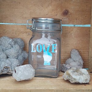 Peut inclure: Un bocal en verre transparent avec un couvercle argenté et un signe de paix bleu et le mot "LOVE" sur le devant. Le bocal est posé sur une surface en bois avec quelques pierres.