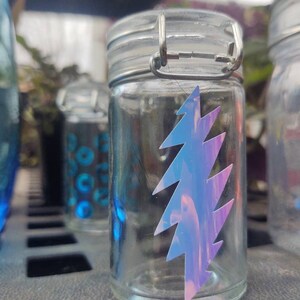 Op de afbeelding: Een heldere glazen pot met een zilveren metalen sluiting en deksel. Een holografische blauwe en paarse bliksemflitssticker is op de voorkant bevestigd. Andere potten zijn zichtbaar op de achtergrond.