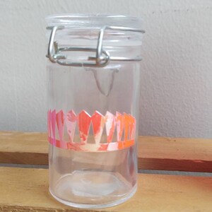 Peut inclure: Un bocal en verre transparent avec une fermeture à fermoir métallique. Le bocal est orné d'un motif géométrique rose et orange au milieu. Le bocal cylindrique semble vide.