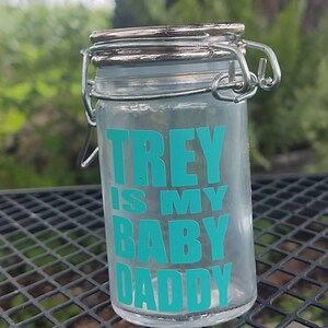 Peut inclure: Un bocal en verre transparent avec un couvercle à charnière et un fermoir en métal. Le bocal porte l'inscription "TREY IS MY BABY DADDY" en lettres turquoise. Le bocal est posé sur une surface métallique noire.