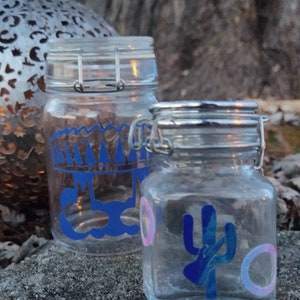 Op de afbeelding: Twee glazen potten met blauwe en roze ontwerpen. De grotere pot heeft een blauw kroon- en wolkenontwerp. De kleinere pot heeft een blauw cactussontwerp. Beide potten hebben zilveren deksels met een sluiting.