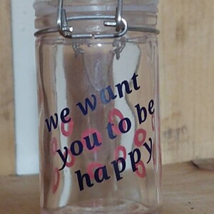 Peut inclure: Un bocal en verre transparent avec un couvercle à charnière et un fermoir en métal. Le bocal porte l'inscription "we want you to be happy" en lettres bleues et roses. Le bocal est posé sur une surface en bois.