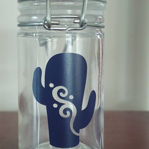 Peut inclure: Un pot en verre transparent avec un couvercle à fermoir métallique. Le pot a un autocollant de cactus bleu avec des volutes blanches.