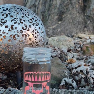 Peut inclure: Un bocal en verre transparent avec un motif rose et blanc en forme de champignon surmonté d'une couronne. Le bocal est posé sur un rocher avec des feuilles et une sphère métallique en arrière-plan.