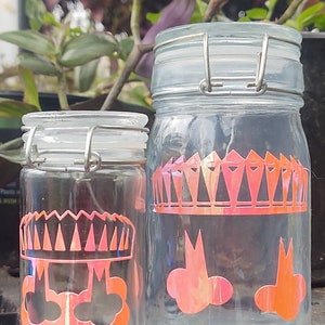 Peut inclure: Deux bocaux en verre avec des couvercles en métal et des motifs de couronne et de nuages orange et noirs. Les bocaux sont transparents et ont une surface texturée.