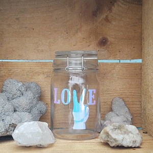 Peut inclure: Un bocal en verre transparent avec un fermoir en métal, orné du mot "LOVE" en lettres irisées et d'un graphisme de signe de la main. Le bocal est entouré de diverses pierres grises et blanches, sur un fond en bois.