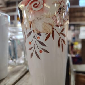 May include: A white ceramic tumbler with a floral design. The top of the tumbler features watercolor-style flowers in shades of pink, peach, and cream. Brown leaves and orange berries adorn the lower portion of the tumbler. The tumbler is cylindrical and sits on a wooden surface.
