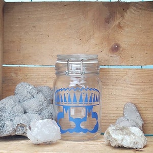 Peut inclure: Un pot en verre transparent avec un motif bleu et blanc d'un crâne portant une couronne. Le pot est posé sur une surface en bois avec quelques rochers.