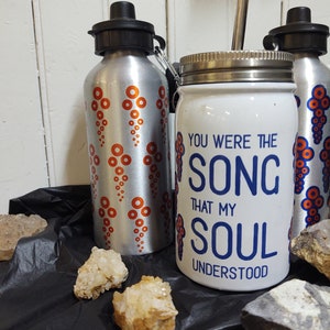 Peut inclure: Deux bouteilles d'eau argentées avec des motifs de points orange et des bouchons noirs sont à côté d'un pot blanc avec le texte "YOU WERE THE SONG THAT MY SOUL UNDERSTOOD" en bleu. Le pot a un couvercle argenté. Des cristaux de quartz sont au premier plan.