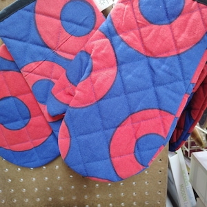 May include: A pair of quilted oven mitts in a vibrant pattern. The mitts feature a blue background with large, overlapping red circles. The edges are trimmed with black binding. The design is a bold, geometric pattern.