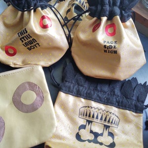 May include: Four gold drawstring bags with black trim and different designs. One bag says "This Still Lawn Boy?" Another says "Page Side High". A third bag has a crown and a fourth bag has a crown with a cloud underneath.