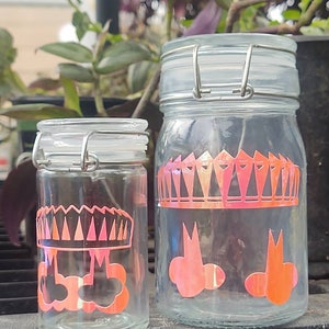 Peut inclure: Deux bocaux en verre transparent avec des couvercles en métal et des motifs géométriques roses et noirs. Le plus grand bocal a un motif de couronne et le plus petit bocal a une couronne et deux nuages.