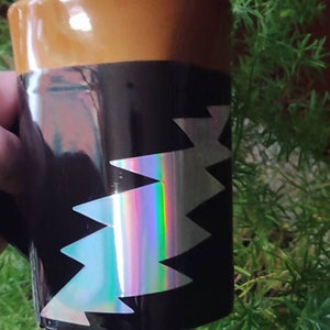 May include: A black coffee mug with a brown rim and a zig-zag design. The zig-zag design has a holographic, iridescent finish. The mug is held in a hand, with green foliage in the background.