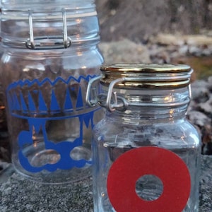 Peut inclure: Deux bocaux en verre avec des couvercles en métal. Le bocal de gauche a un motif bleu et blanc. Le bocal de droite a un cercle rouge avec un cercle plus petit à l'intérieur.