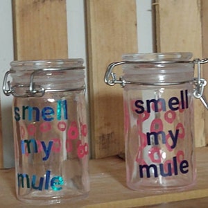 Peut inclure: Deux bocaux en verre transparent avec des couvercles argentés et des lettres roses et bleues qui disent "smell my mule".
