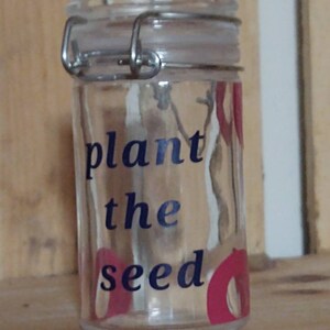 Peut inclure: Un pot en verre transparent avec une fermeture à fermoir métallique. Le pot a un autocollant bleu et blanc qui dit "planter la graine".