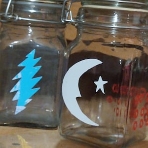 Peut inclure: Deux bocaux en verre avec des couvercles en métal. Le bocal de gauche a un motif d'éclair bleu. Le bocal de droite a un motif de croissant de lune et d'étoile blanc avec du texte rouge.