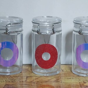Peut inclure: Trois bocaux en verre avec des couvercles en métal et des autocollants circulaires colorés. Les bocaux sont en verre transparent et ont un couvercle en métal argenté avec un loquet. Les autocollants sont un cercle rose et bleu, un cercle rouge et un cercle rose et bleu.