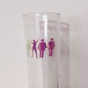May include: A clear glass shot glass with a design of five figures in a rainbow iridescent color. The figures are stylized and appear to be dancing.