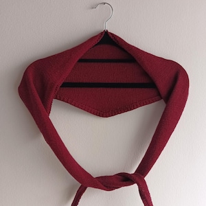 May include: A burgundy-colored knitted shawl with a knotted closure. The shawl is hanging on a black hanger against a white wall. The shawl's design features a triangular shape at the back and long, flowing ties.